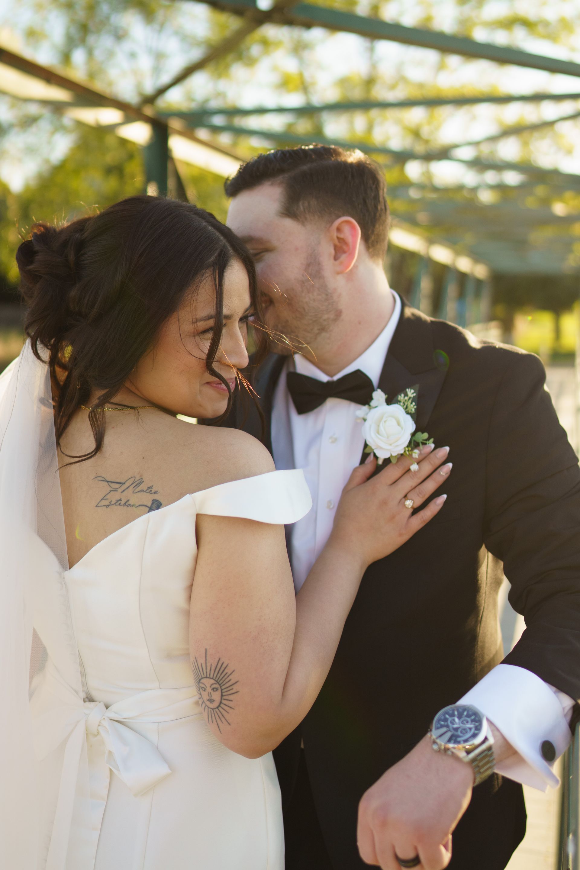 Bride and groom embracing, she smiles at him while he whispers, wearing formal wedding attire; outdoors.