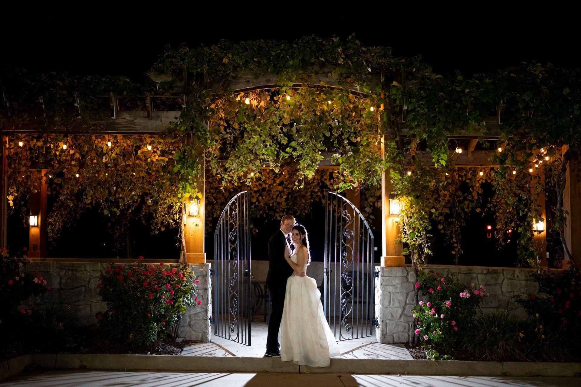 Couple kissing under a vine-covered archway with fairy lights at night.