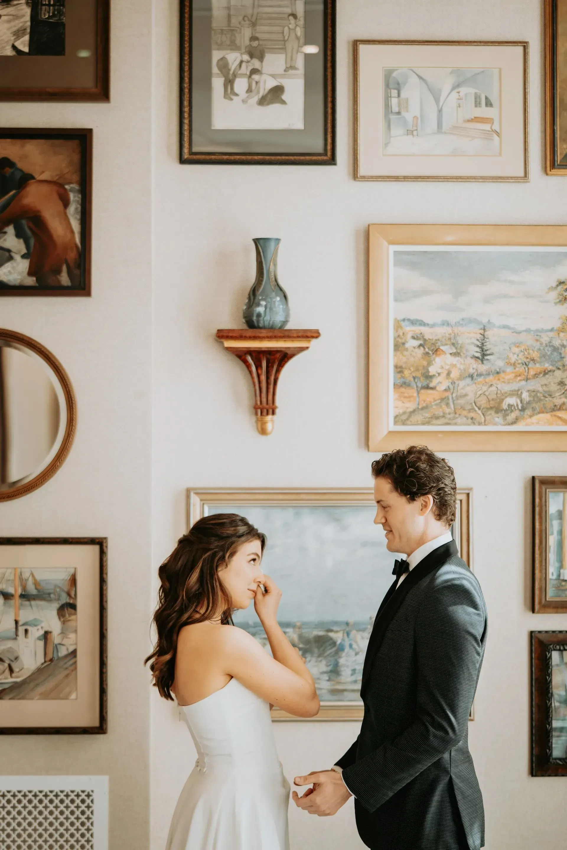 Bride and groom in formal attire, facing each other, holding hands, in front of a wall with framed art.
