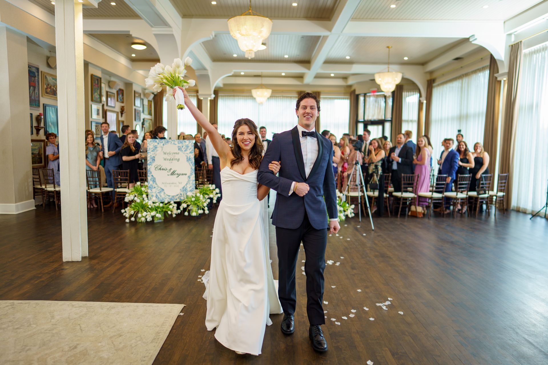 Bride and groom hold hands while crossing a city street; bride in white gown, groom in white jacket, buildings in background.