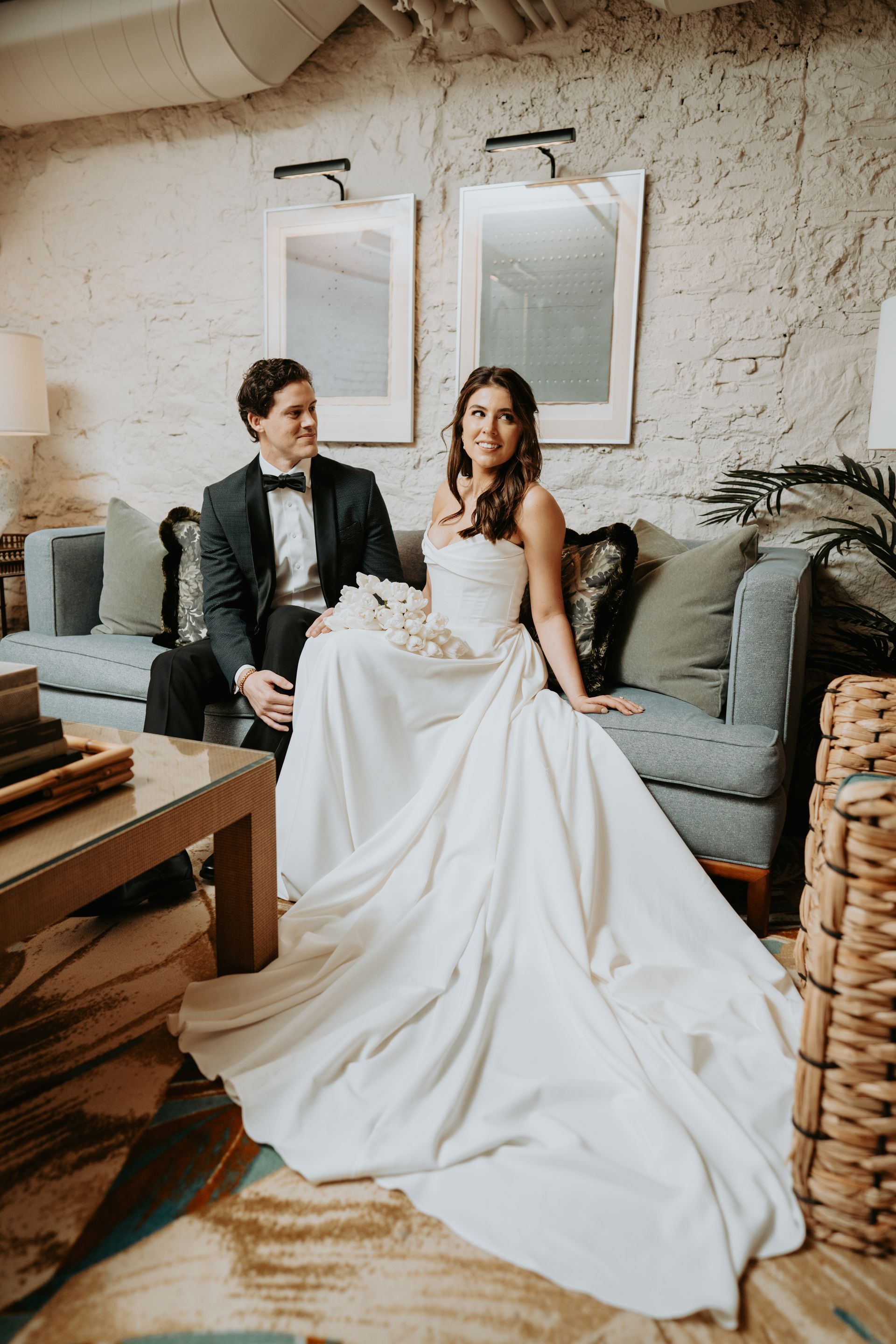 Bride and groom sitting on a blue sofa. Bride in strapless gown, groom in tuxedo, both smiling indoors.