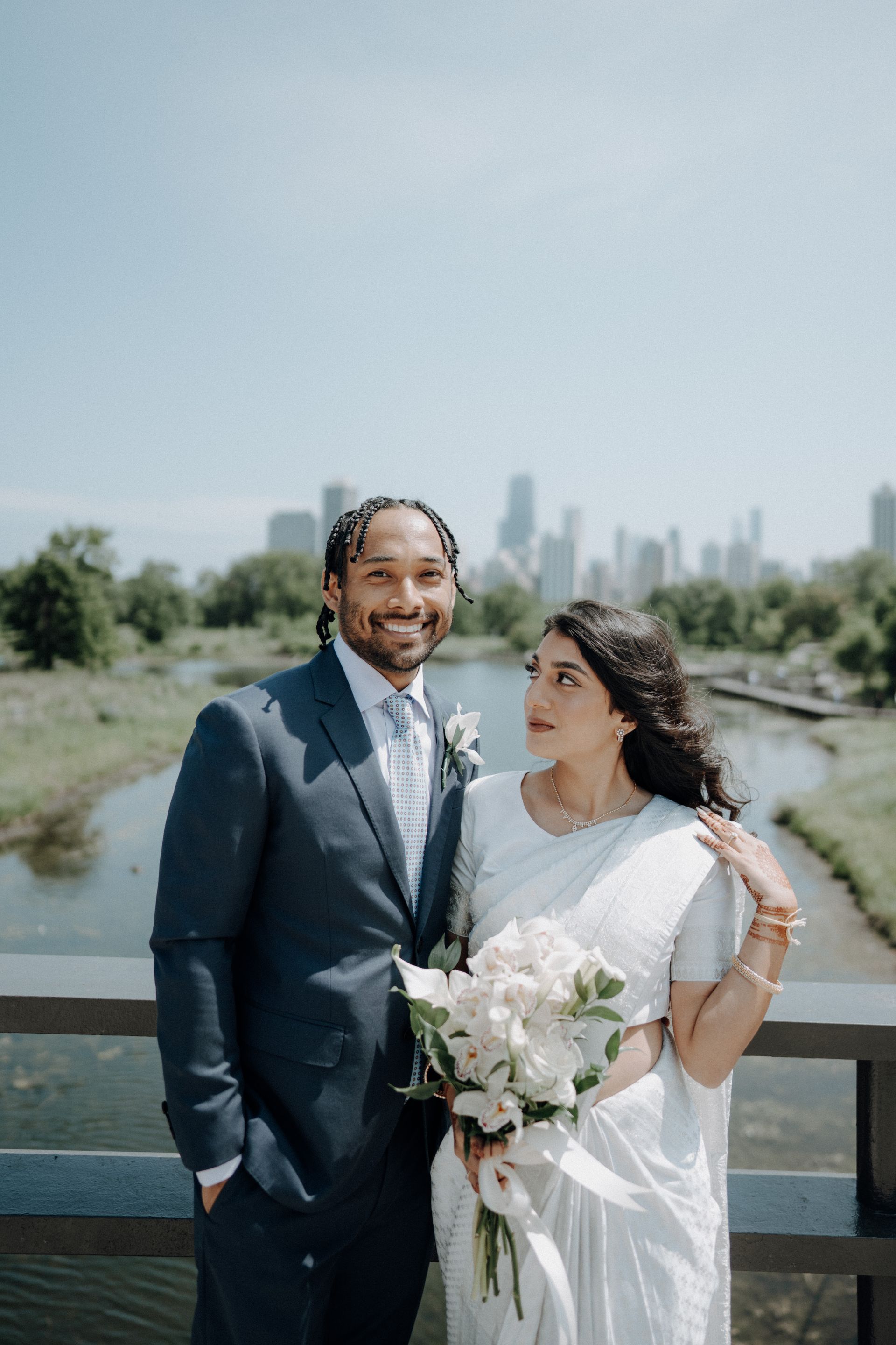 Couple on a bridge smiling, the woman holding a bouquet. City skyline in background.
