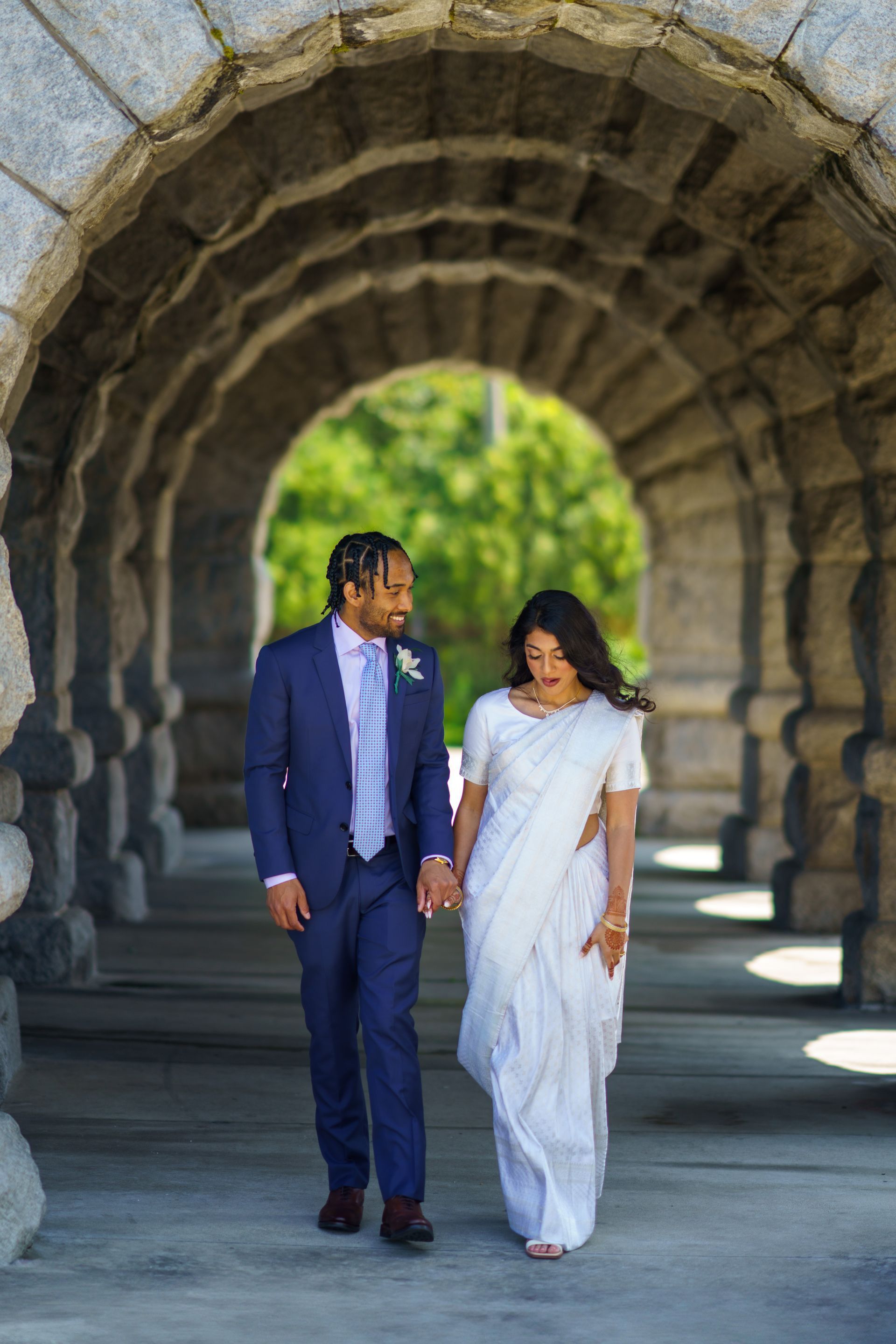 Couple in wedding attire walk through stone archway, holding hands, smiling.