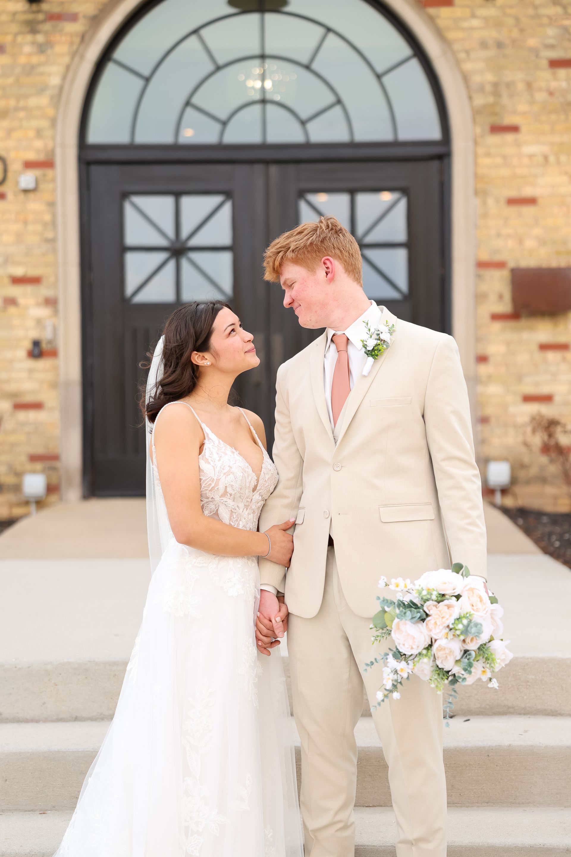Bride and groom holding hands, gazing at each other. Beige suits, white dress, doorway background.