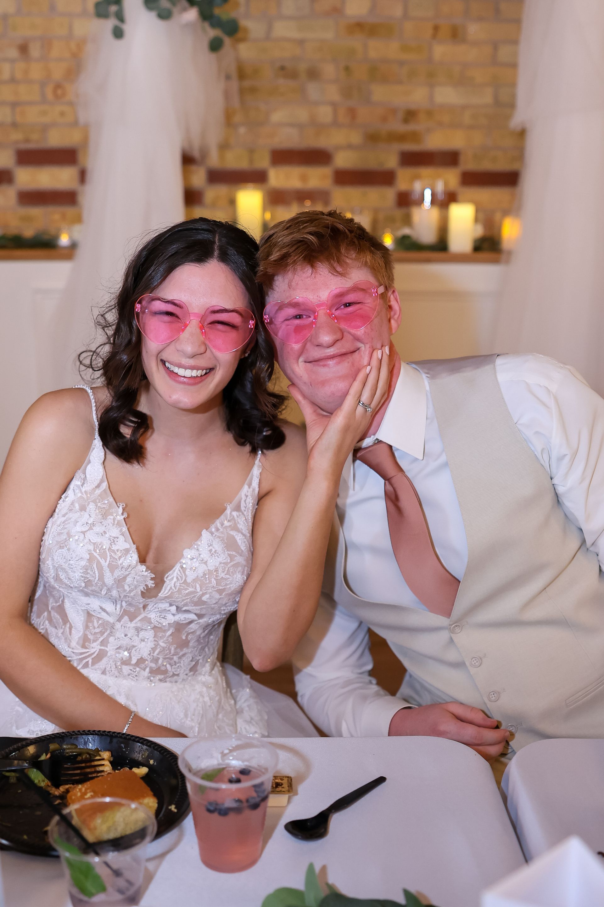 Newly married couple smiles, wearing pink heart-shaped glasses at a wedding reception.