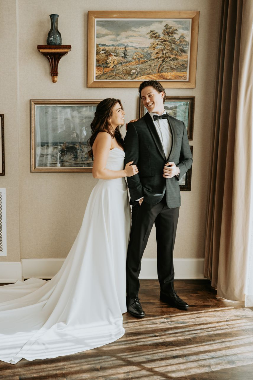 Bride and groom in formal attire, smiling. Interior setting with art and window.