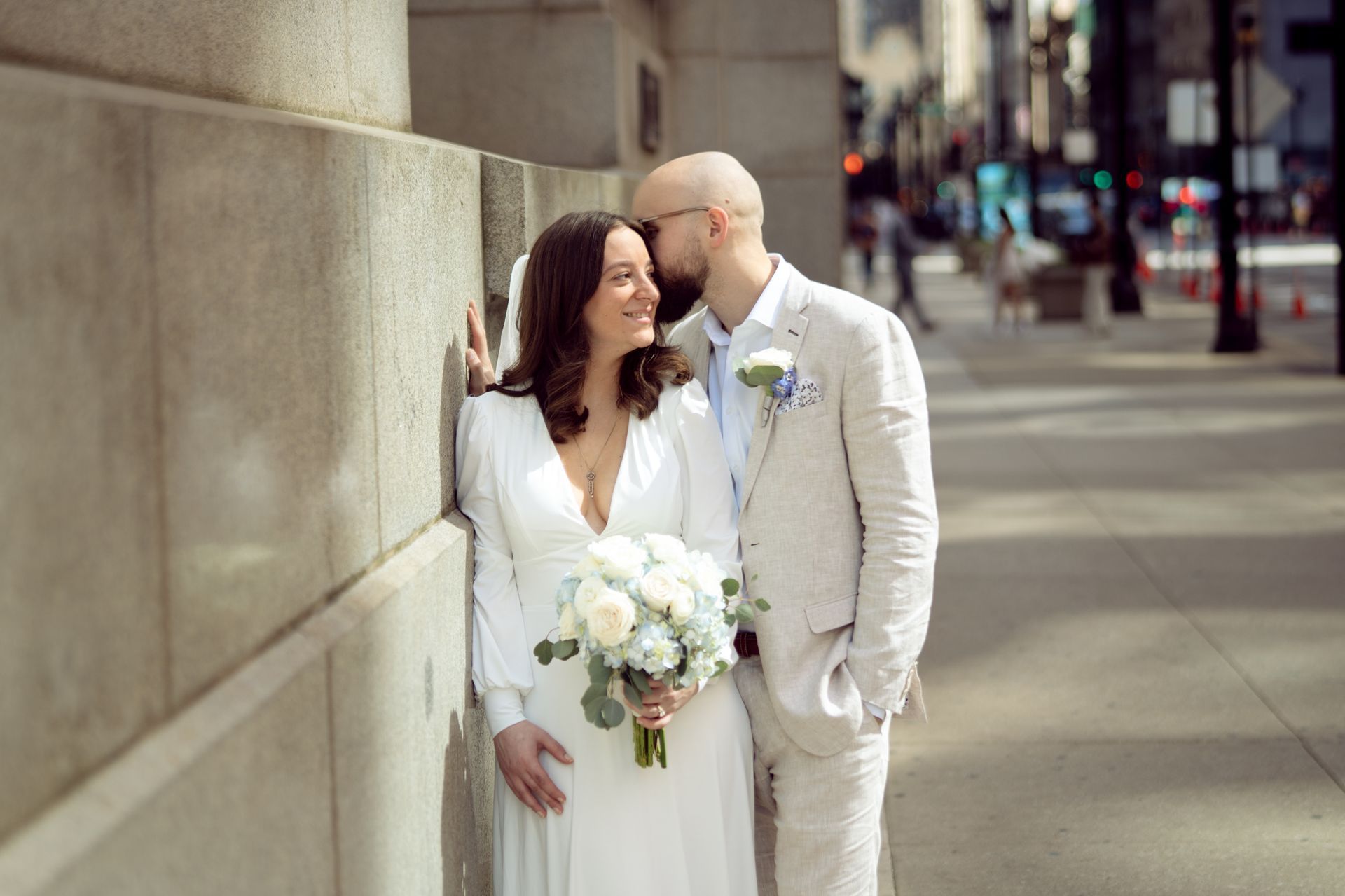 Bride and groom in wedding attire pose by a building. The groom kisses the bride's cheek. They hold a bouquet.