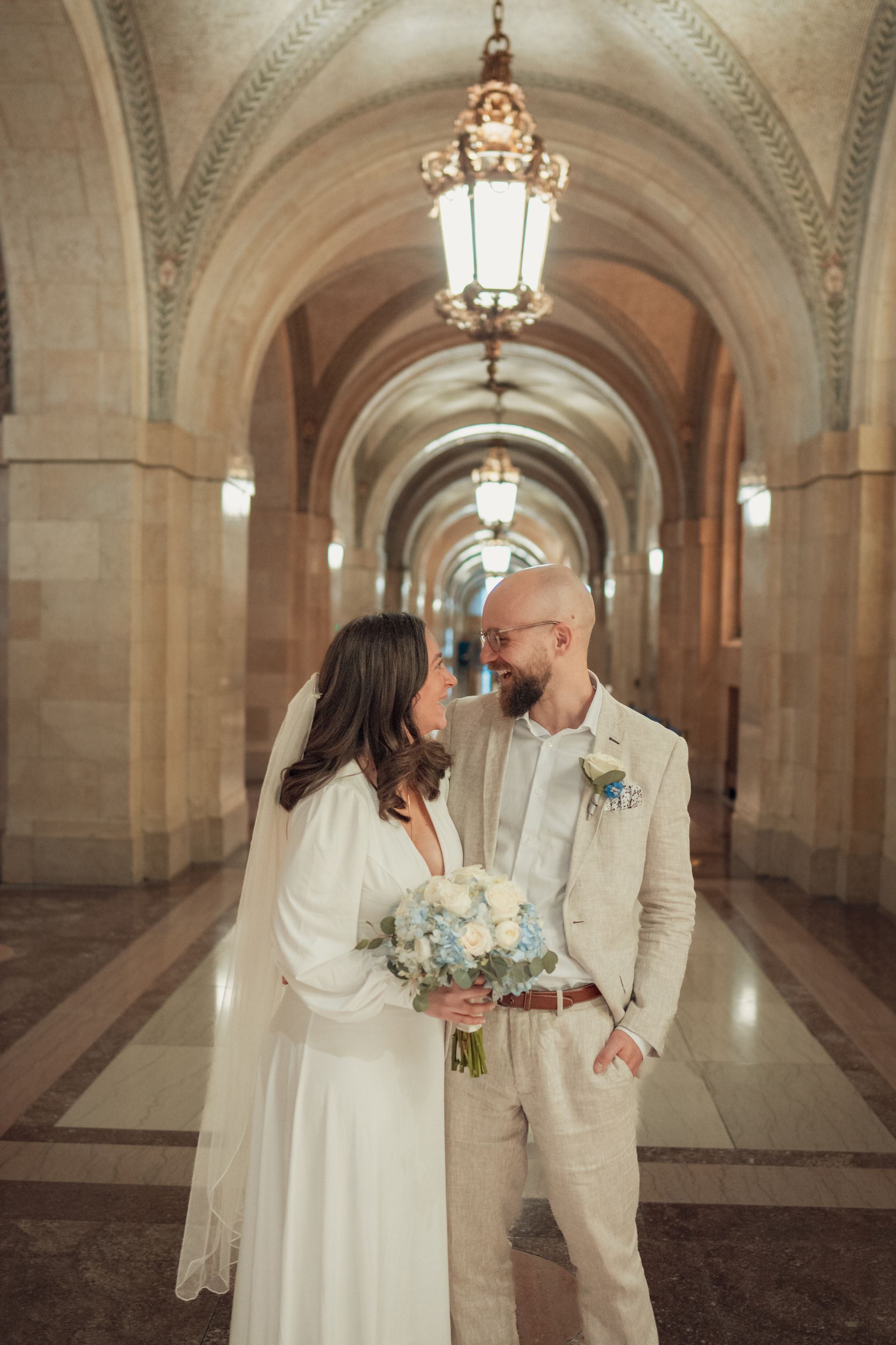 Couple gazing at each other in a stone hallway, holding flowers, before a wedding.