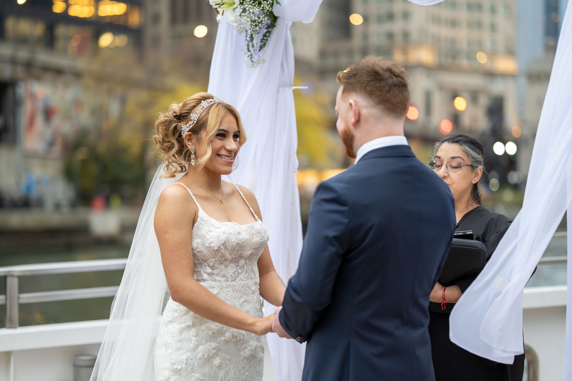 Couple exchanging vows during outdoor wedding ceremony. River and city buildings in the background.