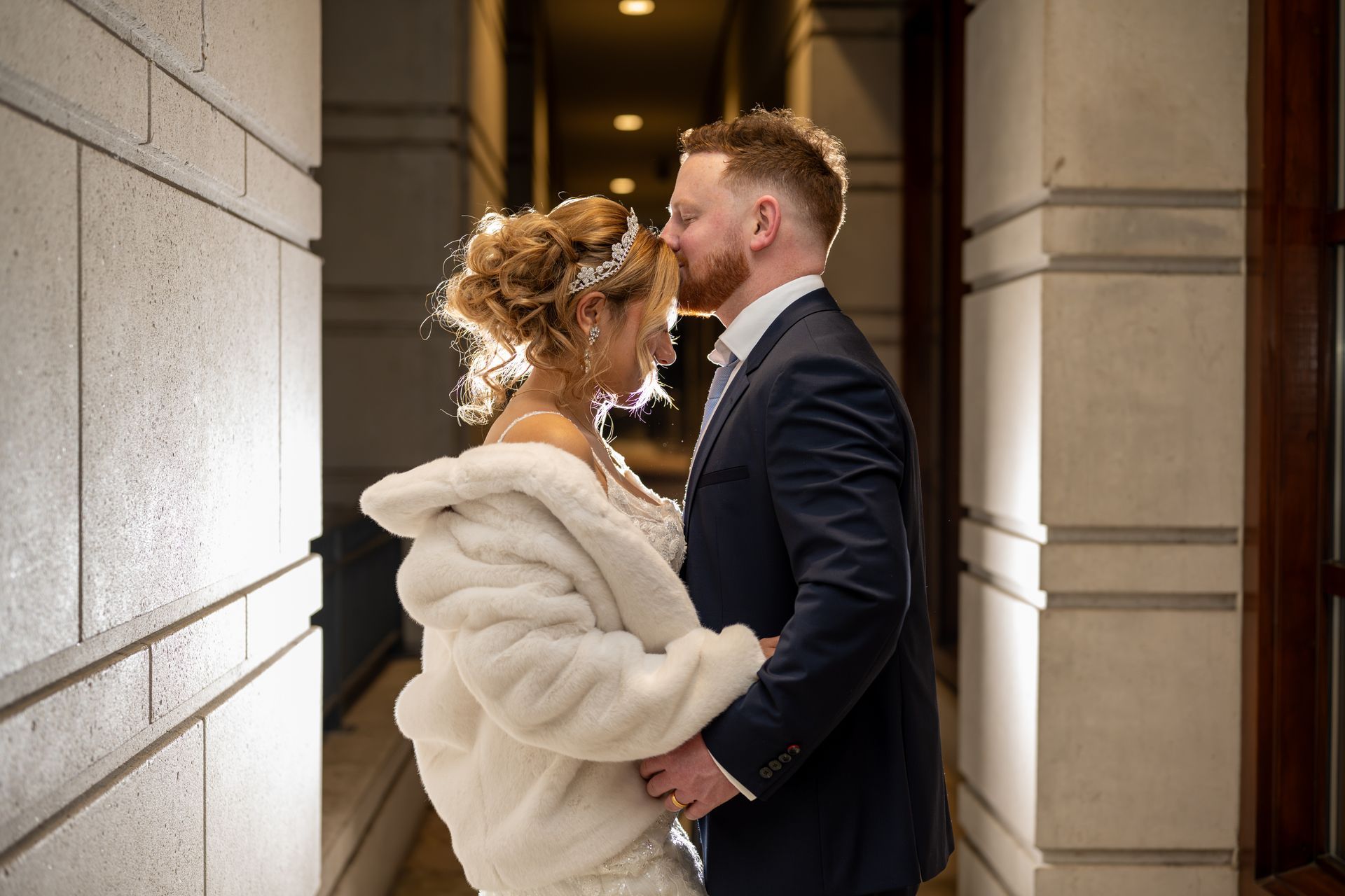 Groom kissing bride’s forehead, both embracing indoors. Bride wears fur shawl, veil. Backlit.