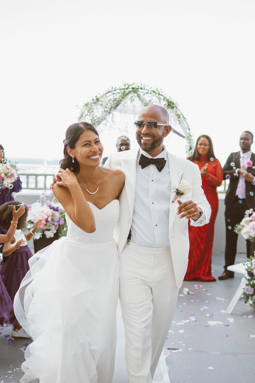 Couple walking, smiling, after a wedding ceremony. Groom in white suit, bride in white dress, outside with floral arch.