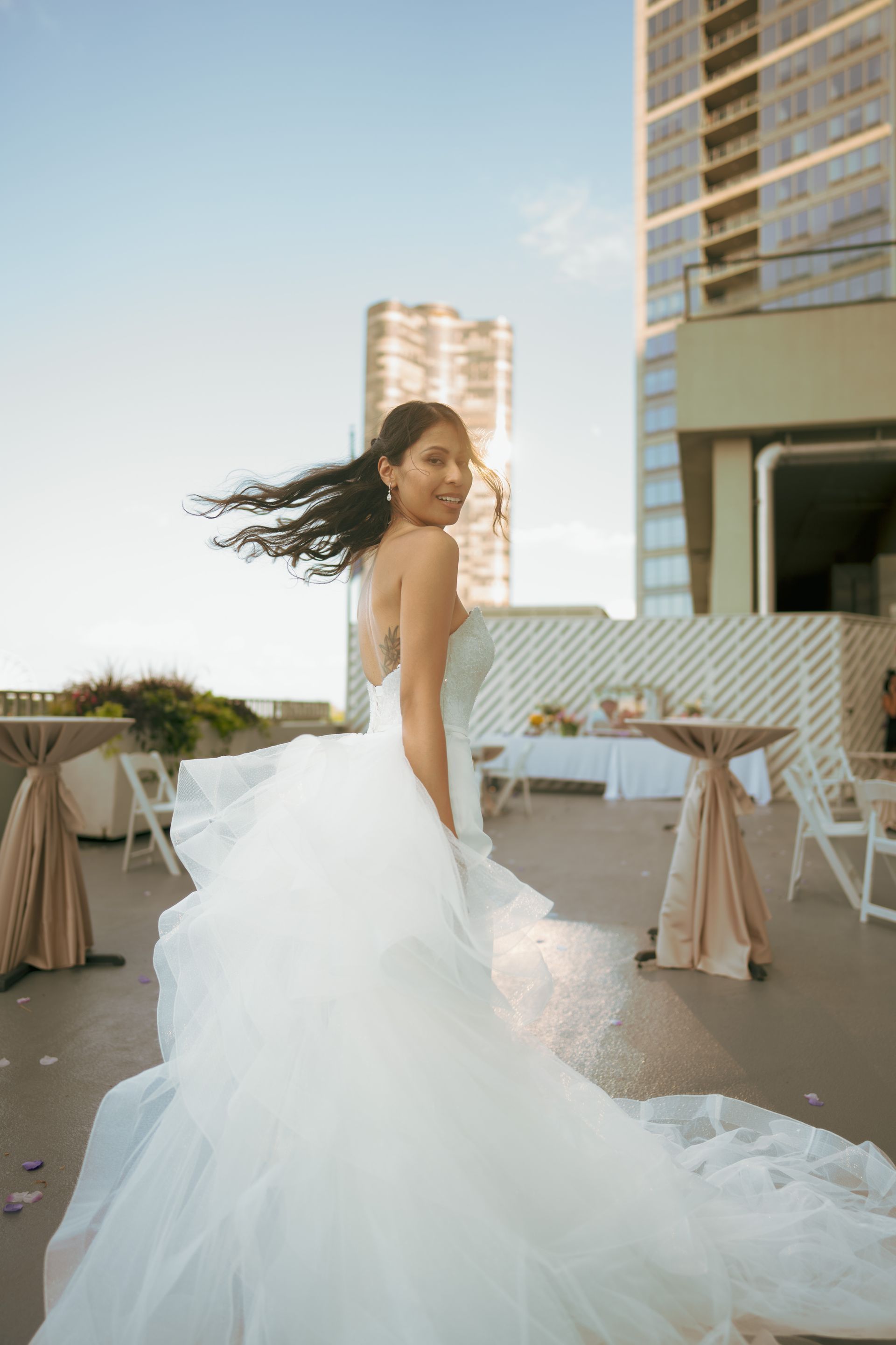 Woman in wedding dress twirling, smiling. Rooftop setting, city buildings in background, sunny.