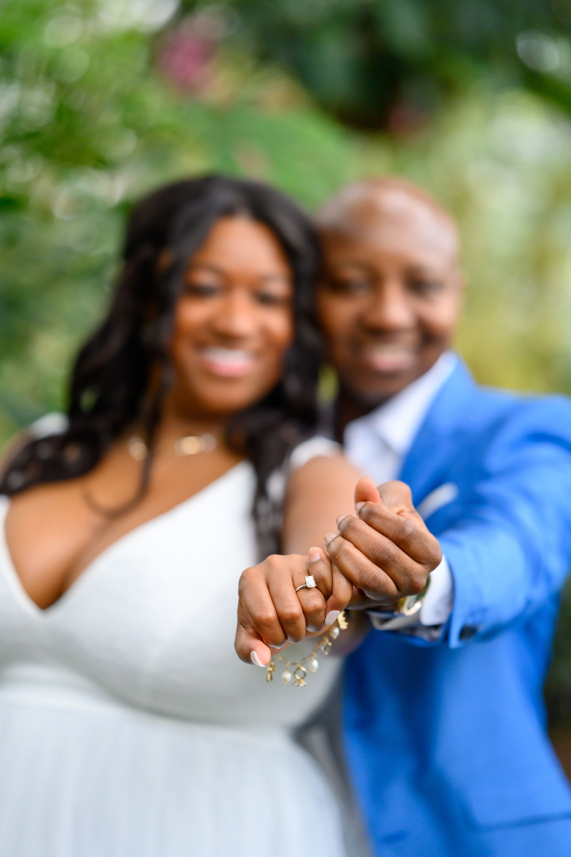 Newlyweds holding hands, showing wedding rings, in front of a blurred green background. One in a blue suit, the other in a white dress.