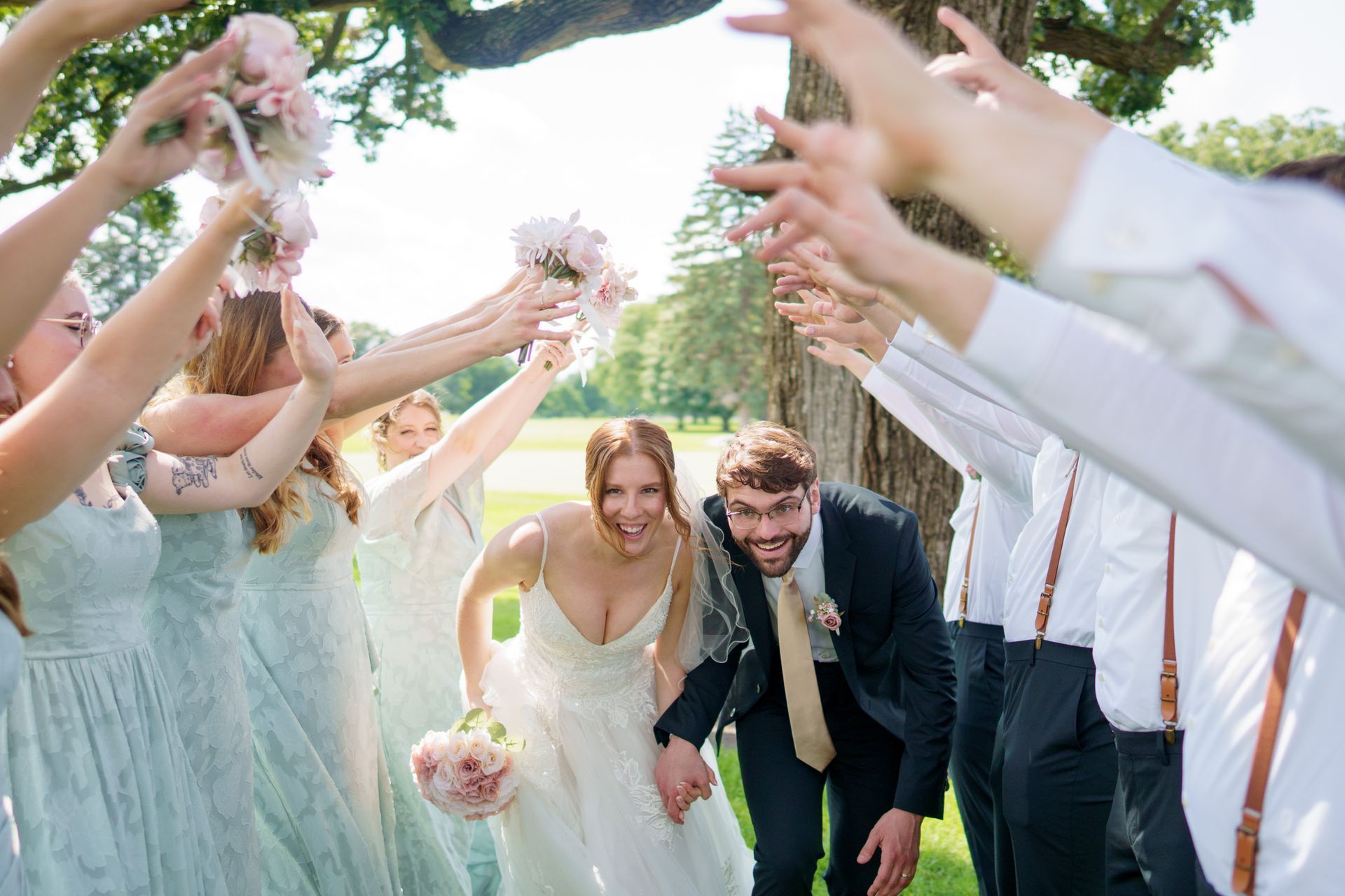 Bride and groom run through a raised arm tunnel by wedding party in a park, smiling with joy.