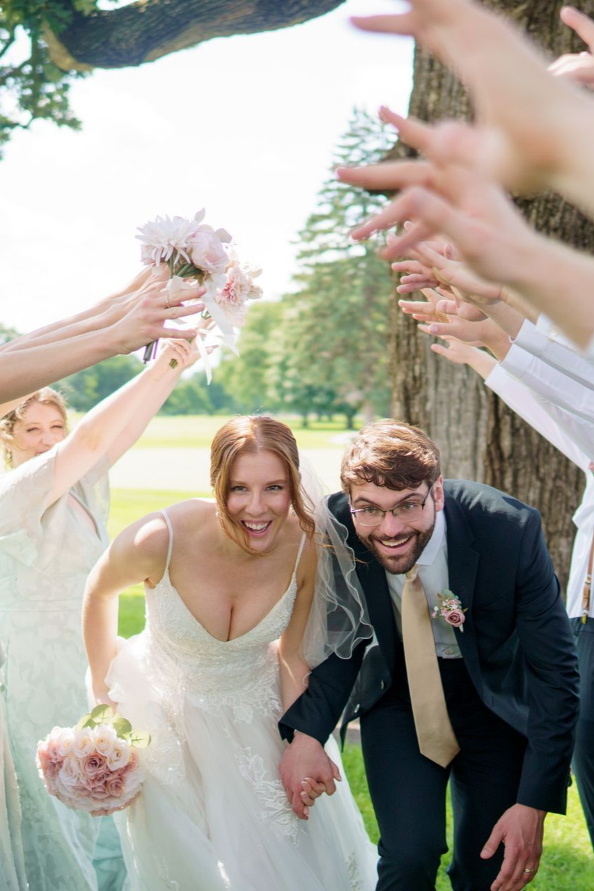 Bride and groom smiling, running through raised arms and bouquets. Outdoors, sunny.