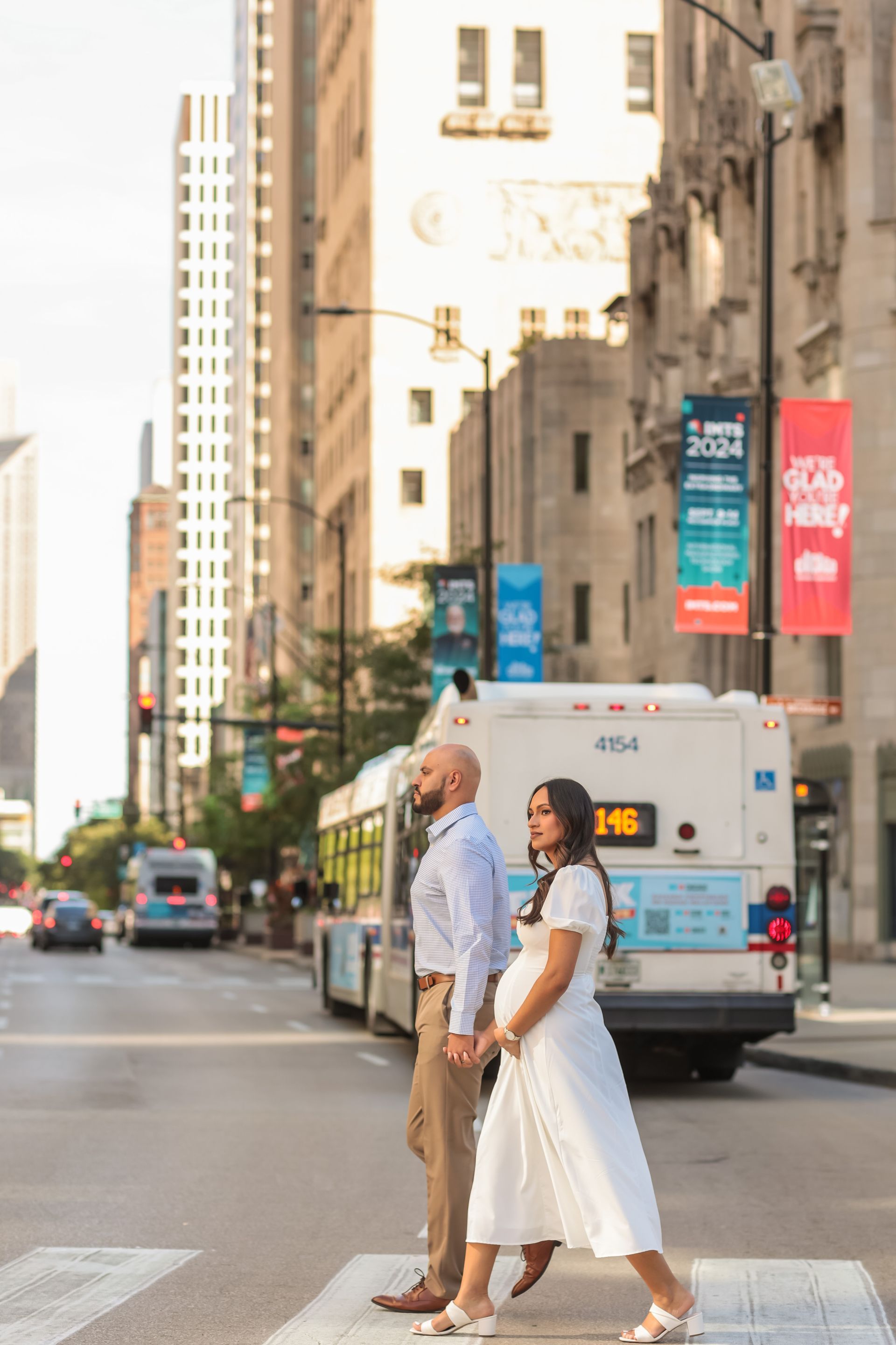 Pregnant person sitting on a stool outdoors, belly exposed, wearing jeans and white shirt, surrounded by trees.