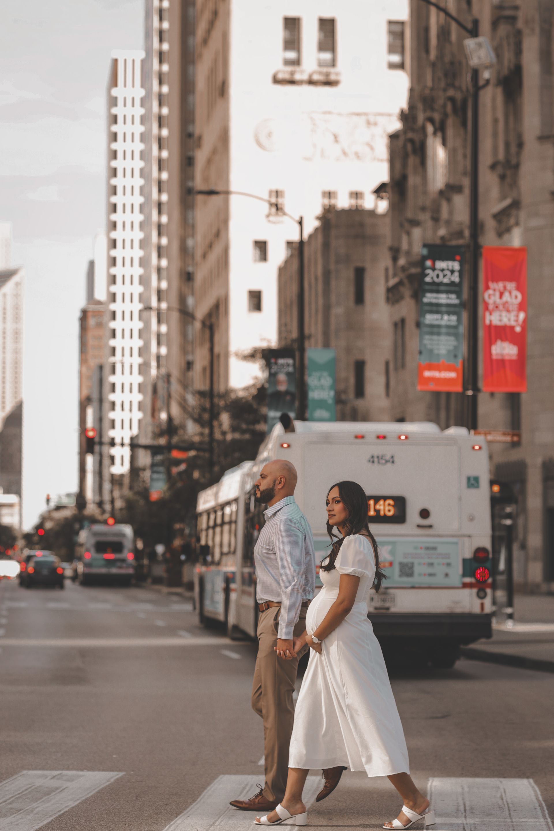 Pregnant woman and partner holding hands, crossing street in city. Bus and buildings in the background.