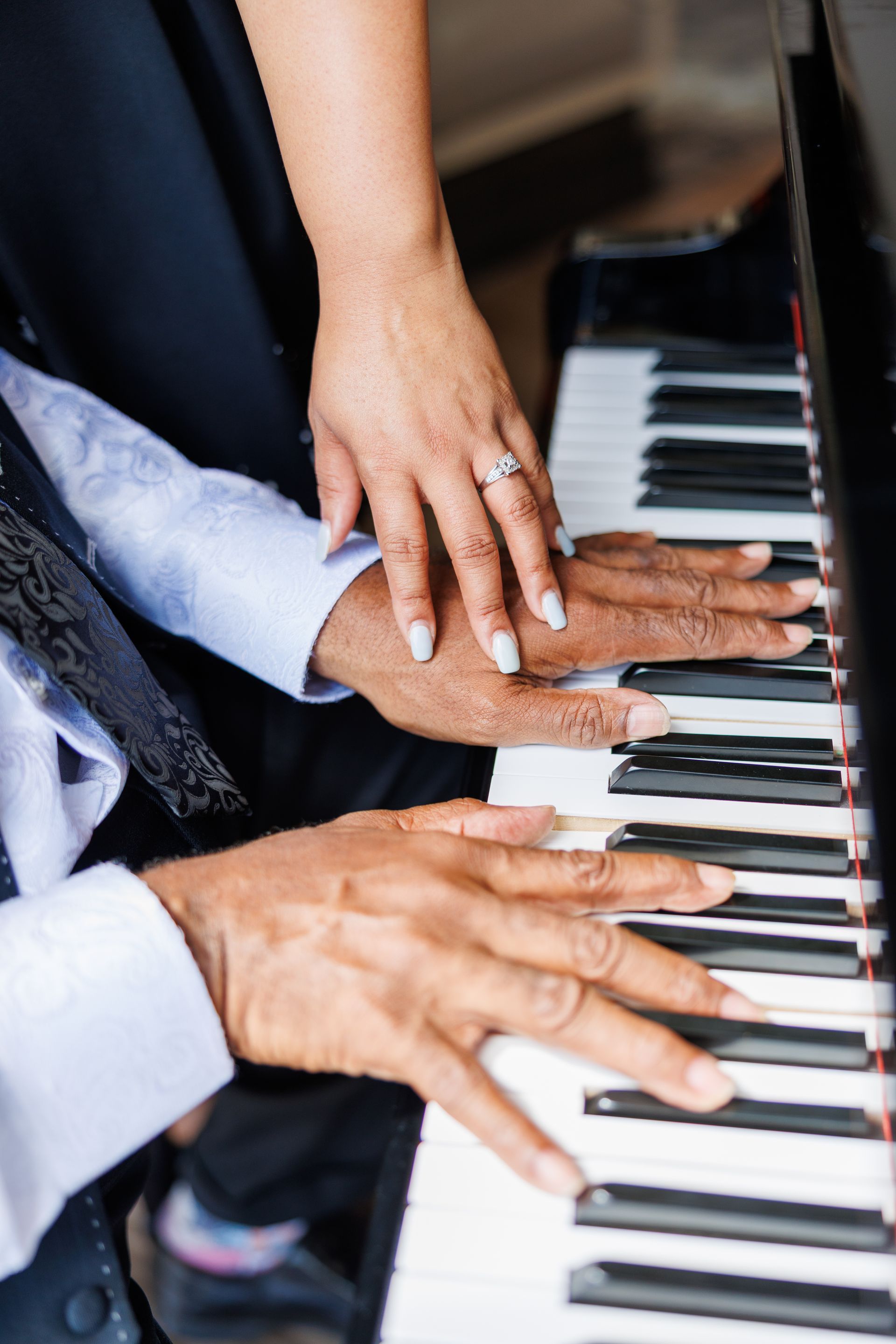 Hands playing piano, another hand rests on top; piano keys are visible.
