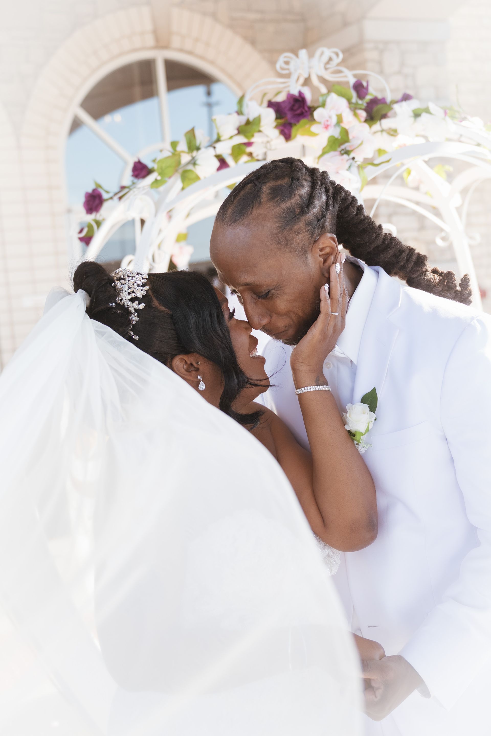 Couple embracing, groom kissing bride's forehead. City setting, sunlight.