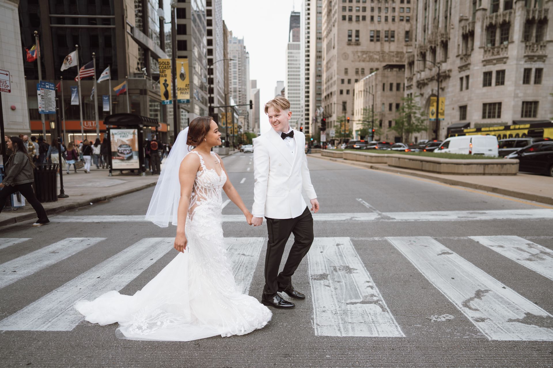 Bride and groom hold hands crossing a city street. Bride wears a white gown, groom a white jacket.