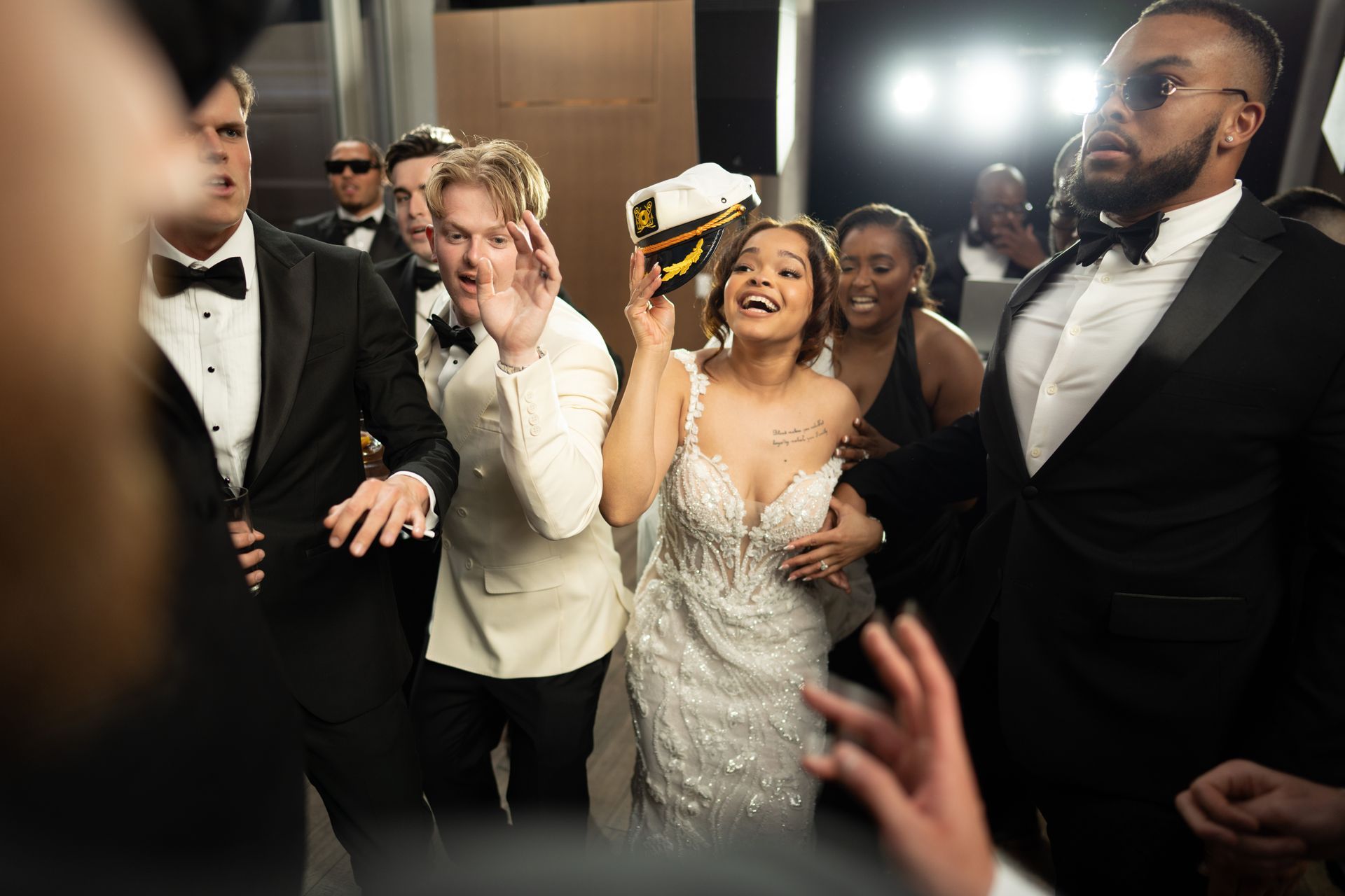 Woman in white dress, holding hat, laughing surrounded by people in tuxedos at event.