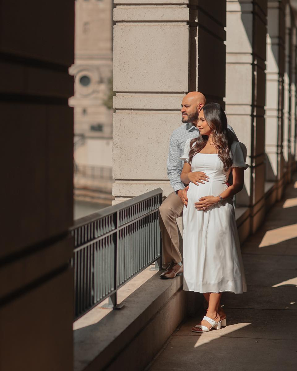 Pregnant woman and partner embrace, looking out from a stone balcony.