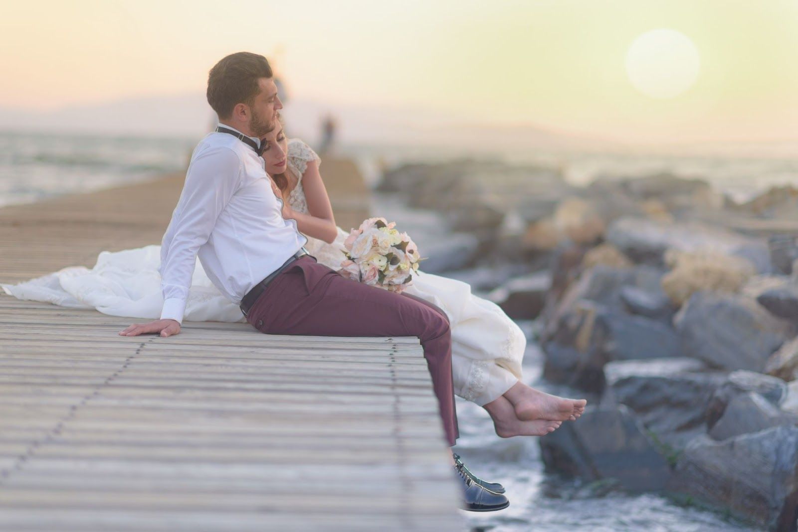 Newlyweds sit on a wooden pier, gazing at the sunset over the water, holding a bouquet.