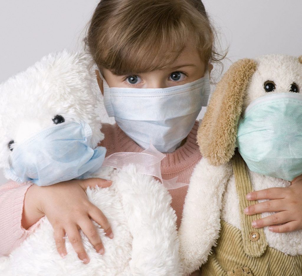 A little girl wearing a mask holds two stuffed animals