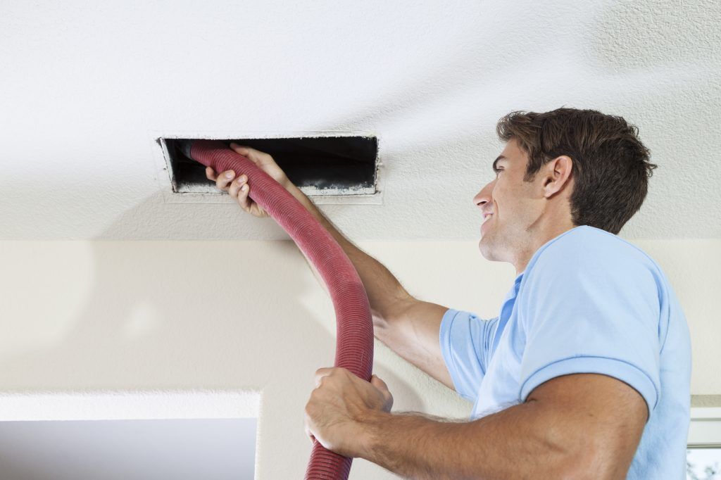 A man is cleaning a ceiling vent with a vacuum cleaner.