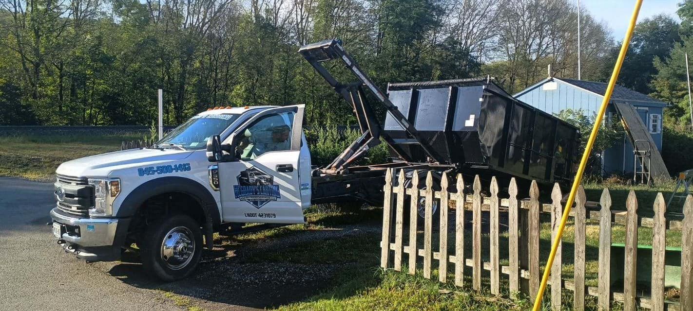 A white roll-off dumpster truck with a black container is parked on a dirt lot next to a wooden fence.