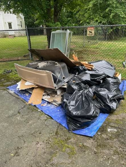 A pile of trash, including black garbage bags, cardboard, and a bathtub, sits on a blue tarp by a chain-link fence.