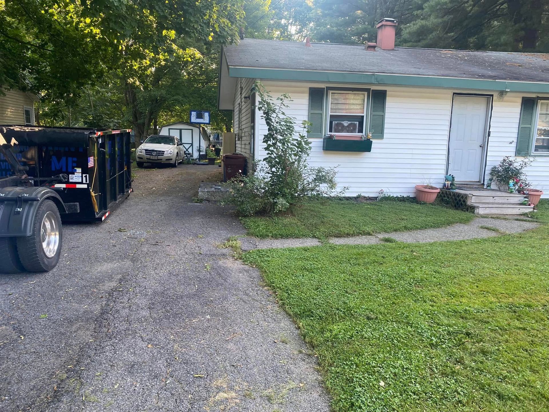 A white, single-story house with a gravel driveway, a black dumpster trailer, and a parked silver car.