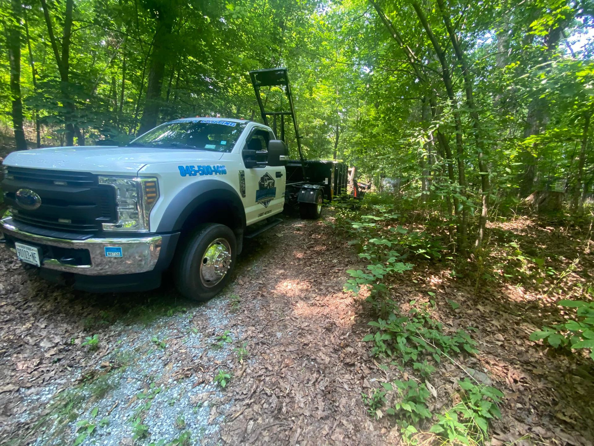 A white service truck with a hydraulic lift parked on a dirt path in a wooded area.