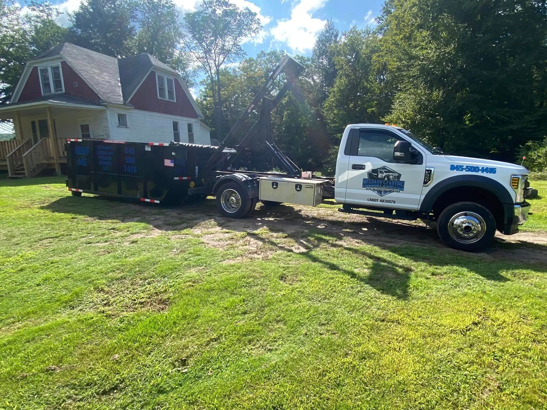 A white roll-off dump truck with a dumpster trailer parked on a grassy lot in front of a house.