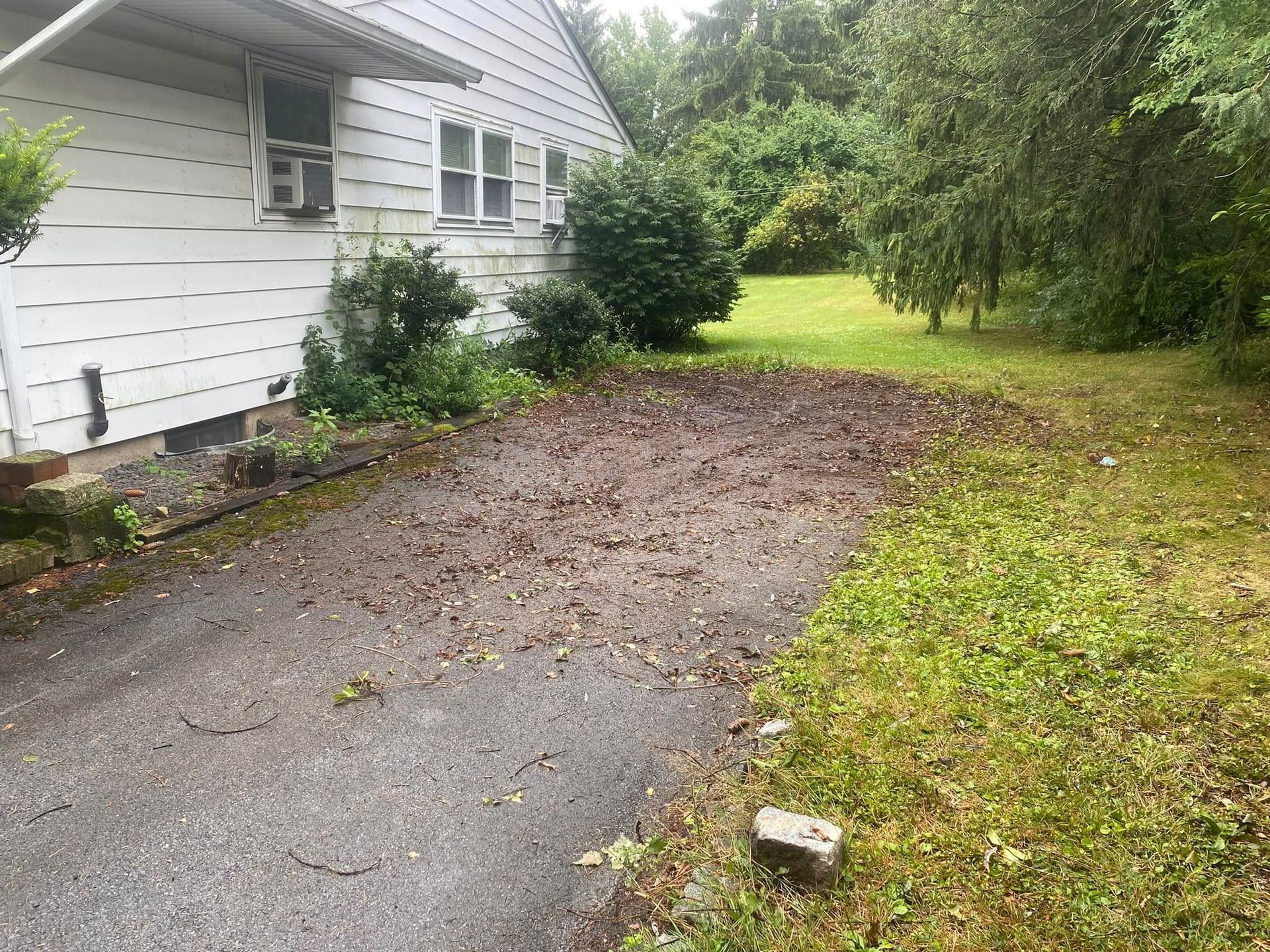 A white house with a side-facing driveway covered in scattered leaves and debris next to a grassy yard.