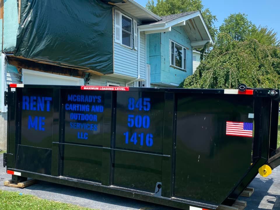 Black dumpster parked in front of a house under construction with blue siding and a dark tarp covering the upper level.