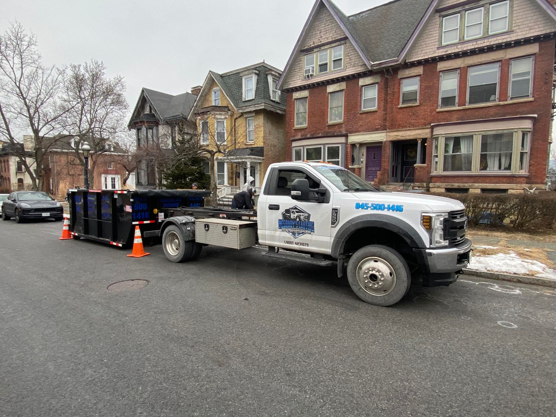 A white dump truck with a blue dumpster attached parked on a city street in front of historic brick houses.