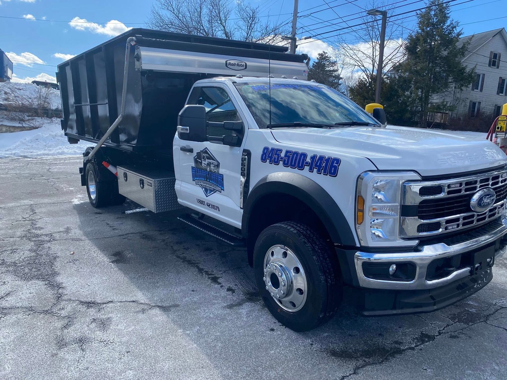 White Ford dump truck parked in a snowy lot, featuring a large black metal container and blue lettering on the door.