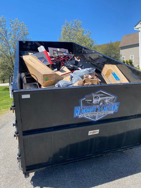 A black dumpster parked on an asphalt driveway is filled with cardboard boxes, debris, and miscellaneous items.
