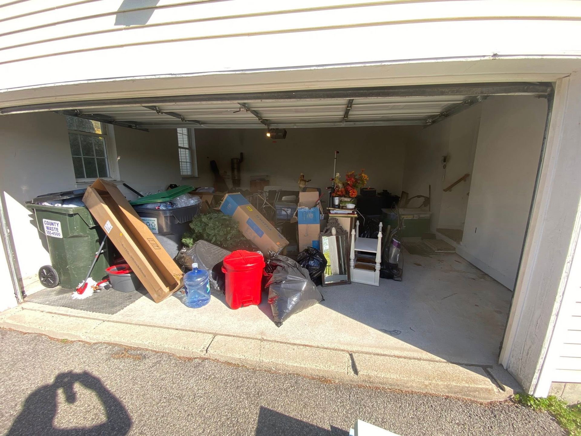A garage with various household items, including a green trash bin, a red container, cardboard boxes, and bags of items.