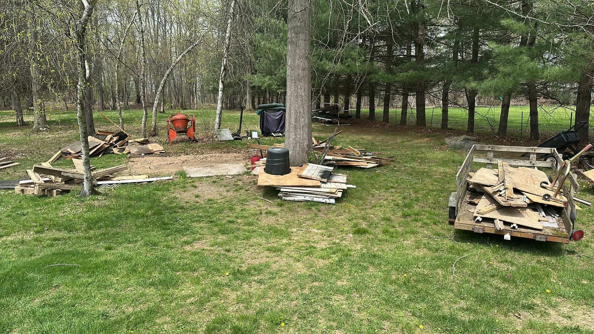 A grassy clearing in a wooded area filled with scattered construction debris, wood planks, and a trailer loaded with trash.