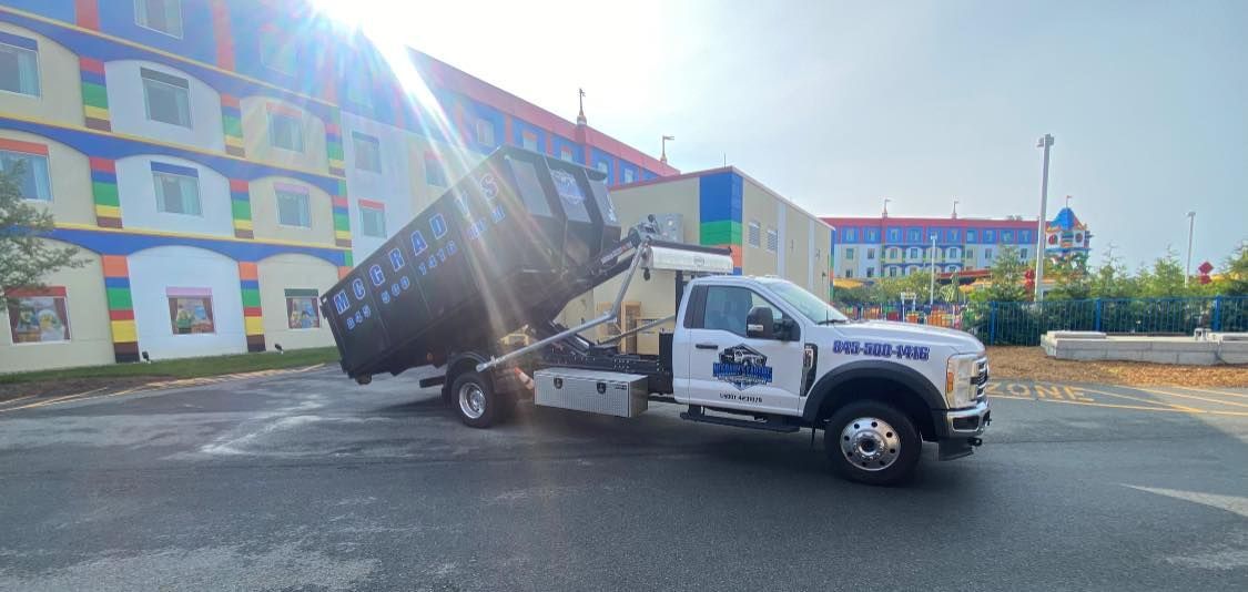 A white roll-off dumpster truck is tilting its large black container to unload it in a paved lot near a colorful building.