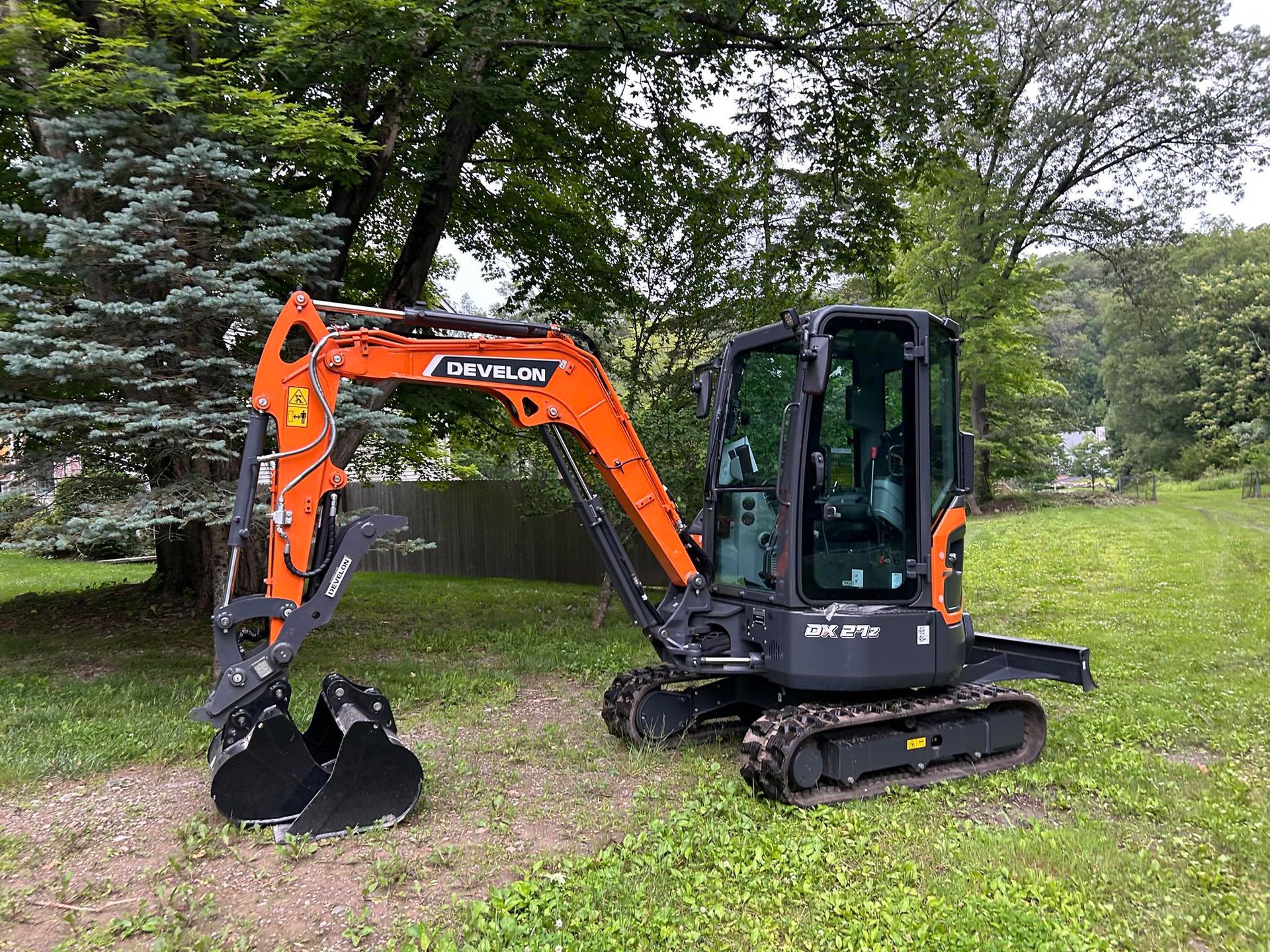 An orange and dark gray compact excavator sits on a grassy field in front of green trees.