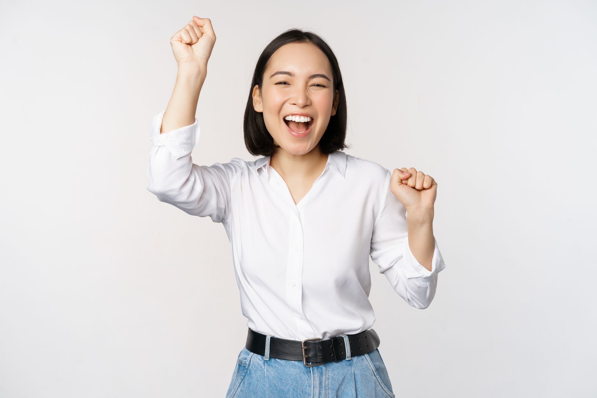 Woman with short dark hair, smiling and raising fists in the air, white shirt, light background.