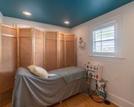 Small room with a massage table, folding privacy screens, white walls, teal ceiling, and a window