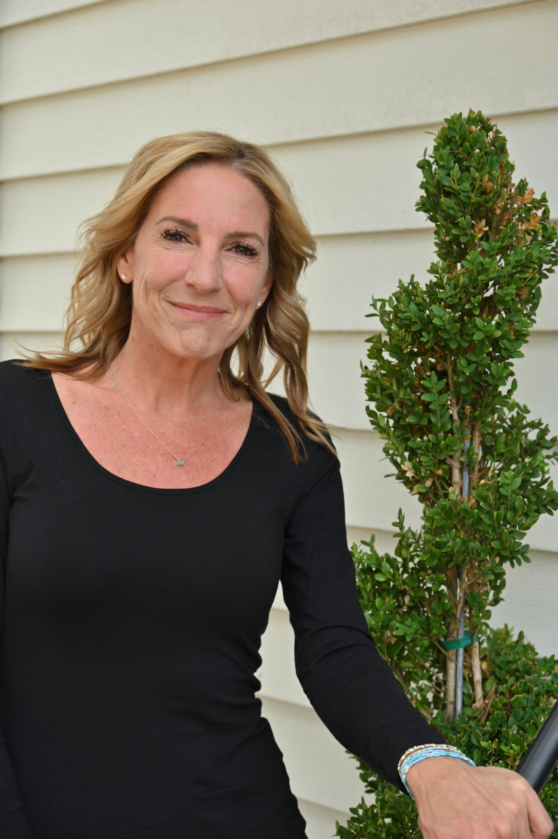 Woman in a black top smiling outdoors beside a trimmed evergreen shrub