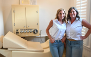 Two women standing beside a cream-colored medical examination chair in a clinical room