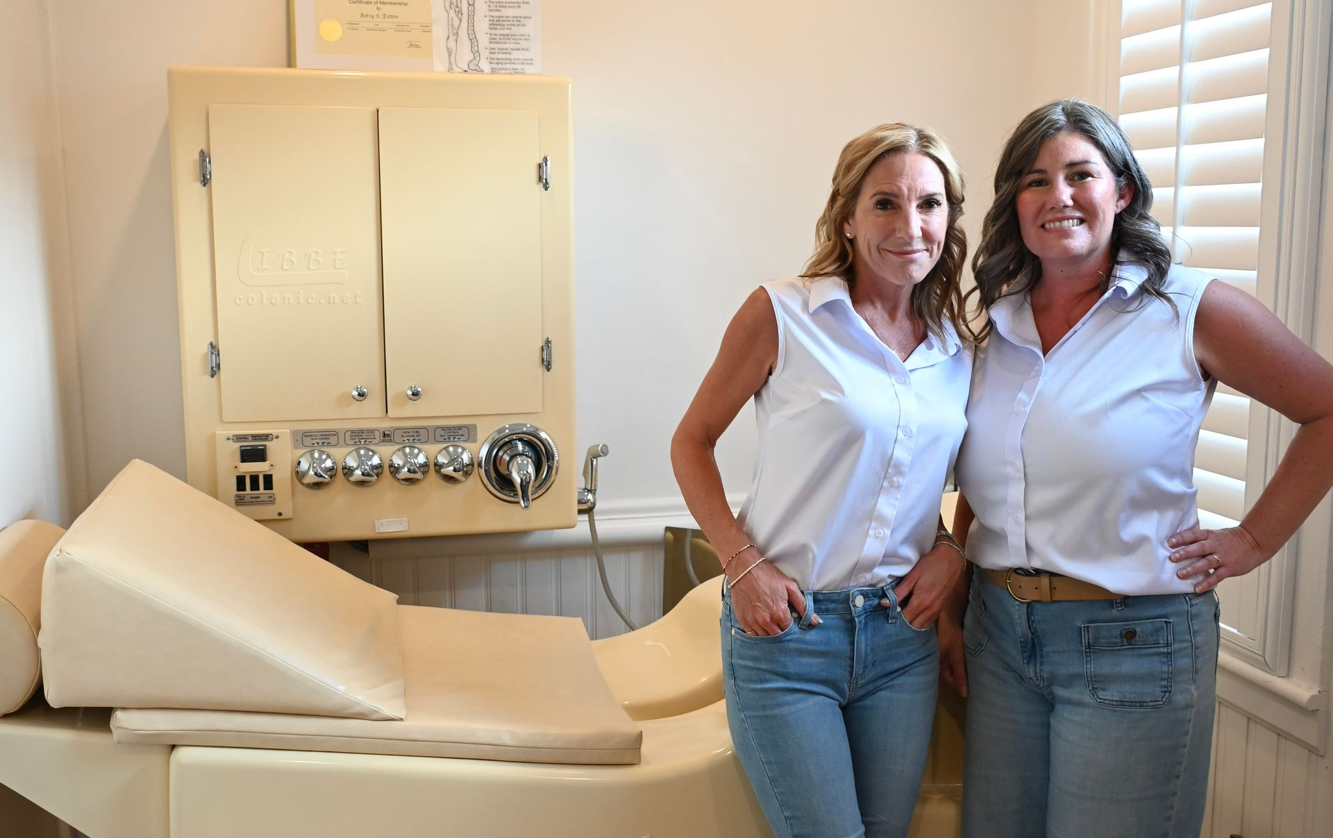 Two women standing beside a cream-colored medical examination chair in a clinical room