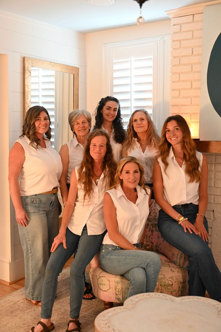 Six people in white tops and jeans posing together in a bright room with windows and a brick fireplace