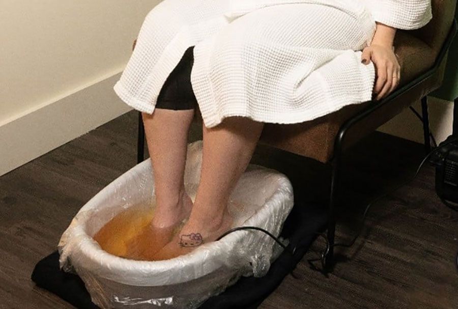 Person in a white robe soaking feet in a plastic foot bath on a chair.